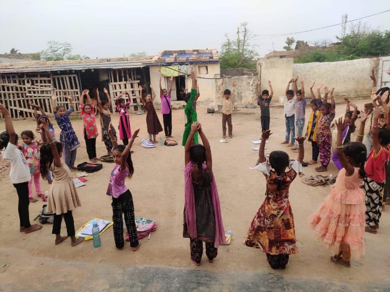 Village children standing in a courtyard with their arms raised, exercise books on the ground