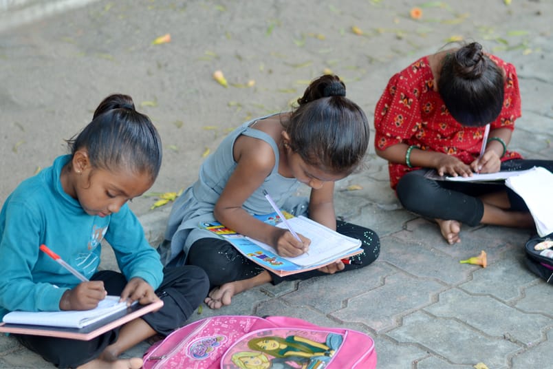 Three girls sitting on paving in the shade, exercise books open on their knees, writing with school bags beside them