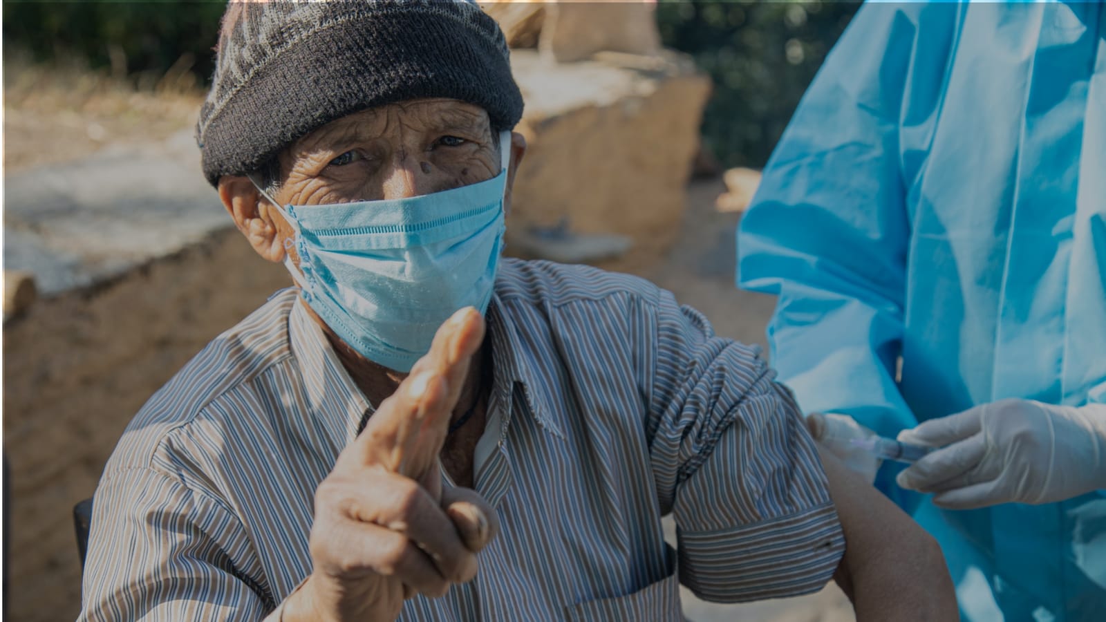 An older man in a woollen cap and surgical mask holding up a hand while a health worker in a blue protective gown gives him an injection in the arm, outdoors beside a mud-walled house